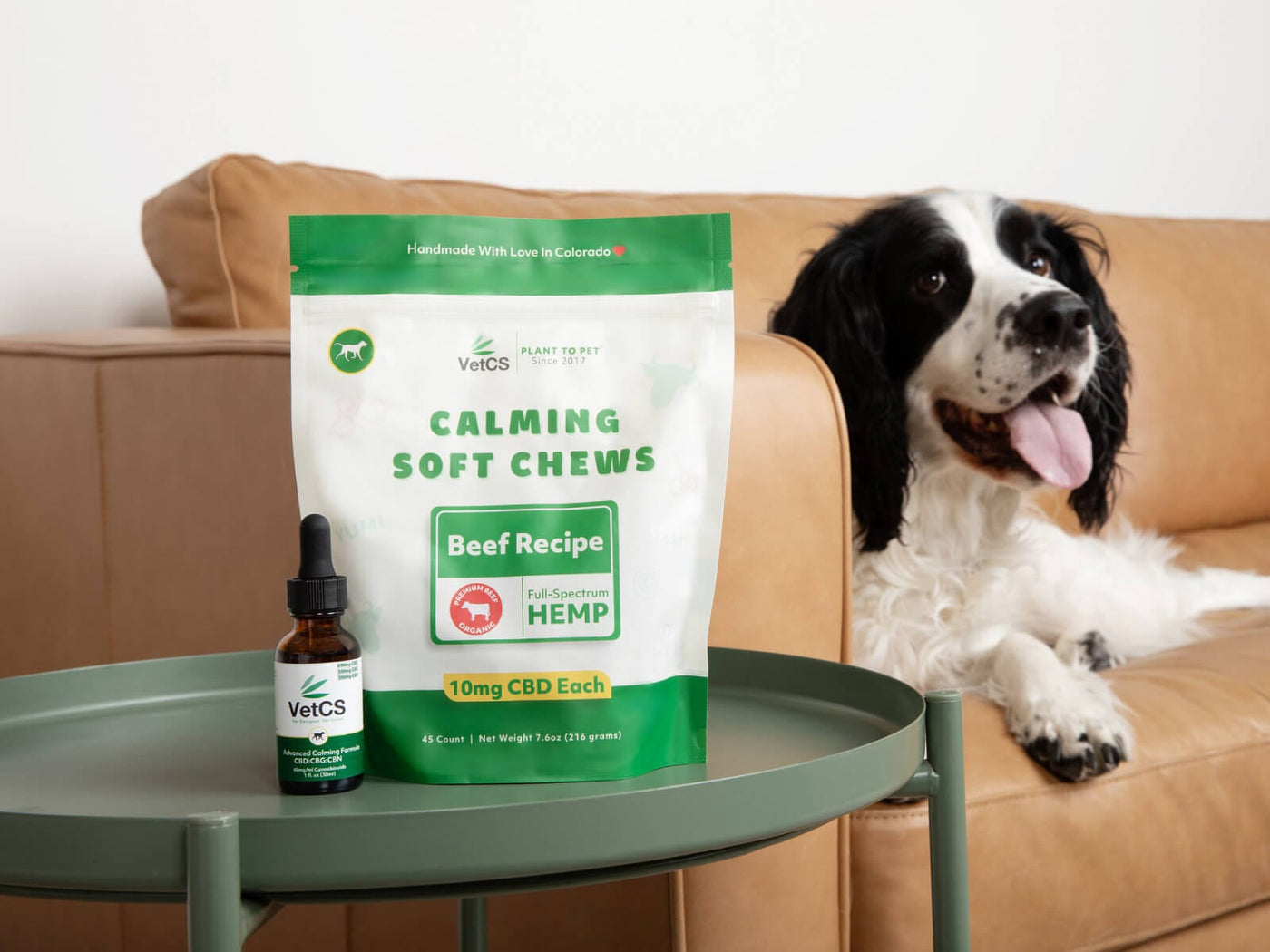 Dog sitting on a couch next to a package of 'Calming Soft Chews' with a tincture on a table