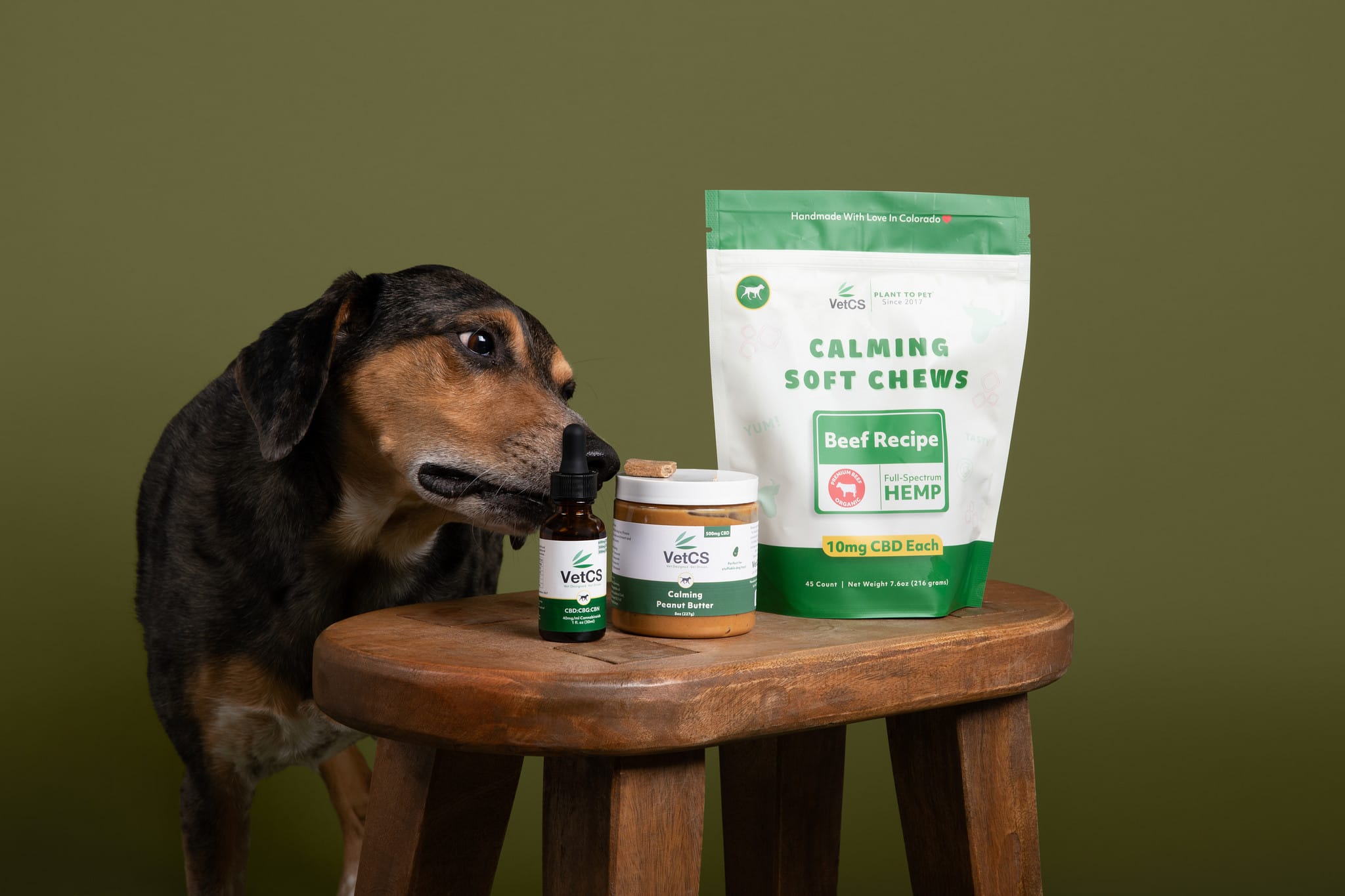 Brown and black dog sniffing VetCS calming soft chews, calming peanut butter, and advanced calming formula with  green background